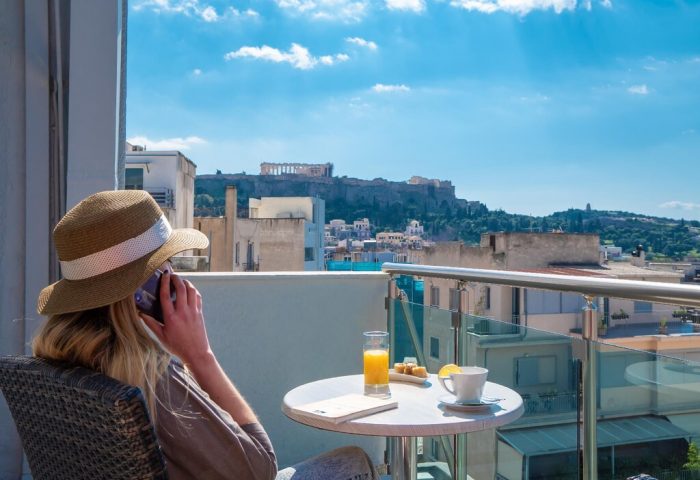 Woman on phone at Attalos Hotel, a gay-friendly hotel, with Acropolis view. Enjoying breakfast on balcony in Athens. Travel advice for gay men.
