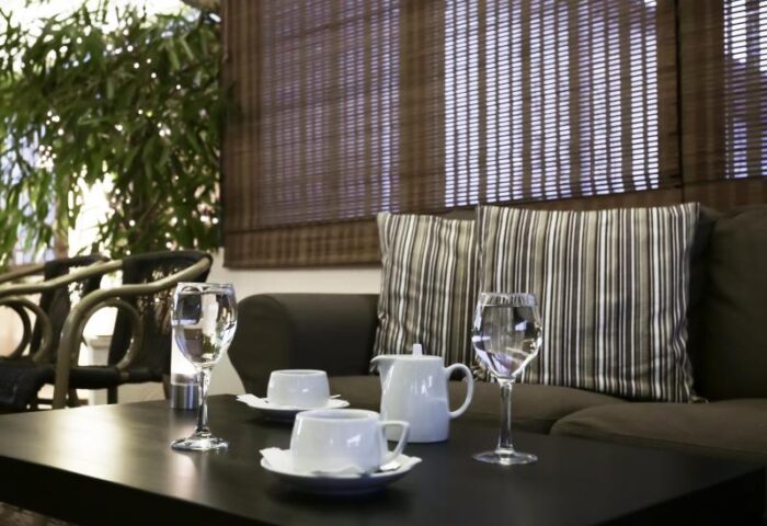 Attalos Hotel's lounge area, a gay-friendly hotel. Table with tea set and glasses, sofa with striped cushions, and bamboo blinds. Relaxing space.
