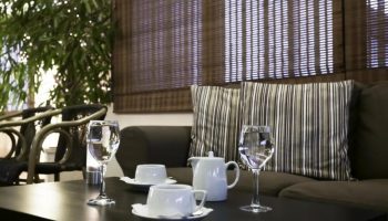 Attalos Hotel's lounge area, a gay-friendly hotel. Table with tea set and glasses, sofa with striped cushions, and bamboo blinds. Relaxing space.