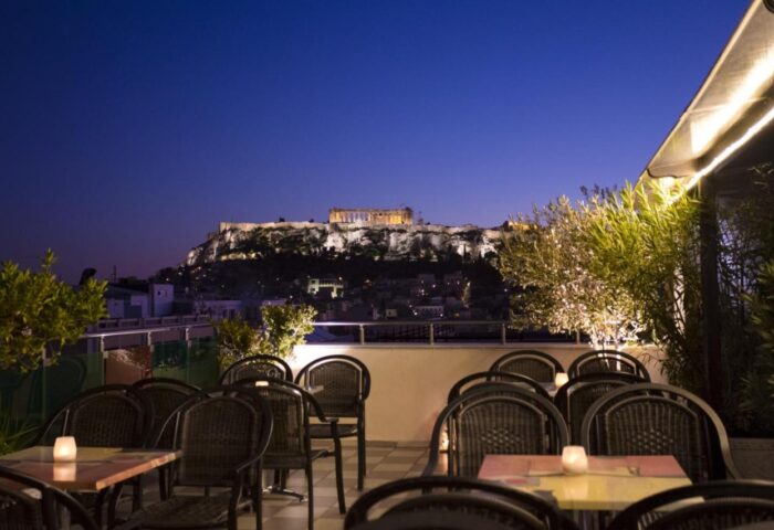 Attalos Hotel's rooftop bar at night. The gay-friendly hotel offers views of the Acropolis in Athens, Greece. Tables and chairs visible.