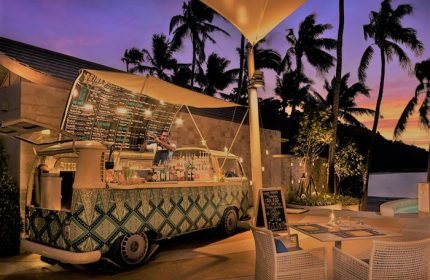 Avani+ Samui Resort's gay-friendly bar. A bartender mixes drinks in a converted VW van bar. Tables and chairs sit nearby under palm trees.