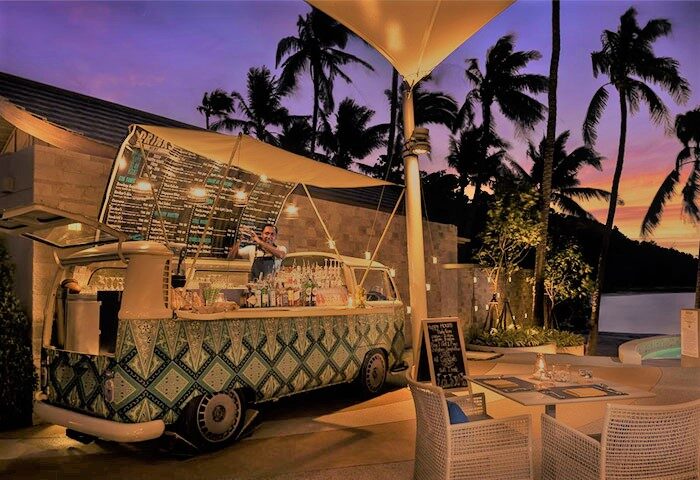 Avani+ Samui Resort's gay-friendly bar. A bartender mixes drinks in a converted VW van bar. Tables and chairs sit nearby under palm trees.