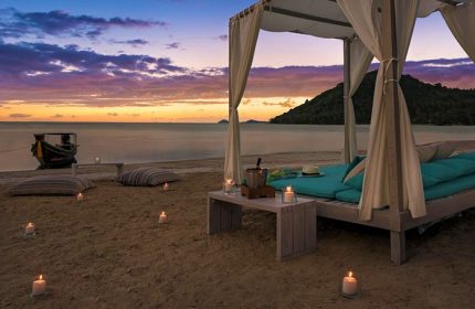 Beachfront canopy bed at sunset, Avani+ Samui Resort, a gay-friendly hotel. Candles illuminate the sand near a boat. Romantic getaway.