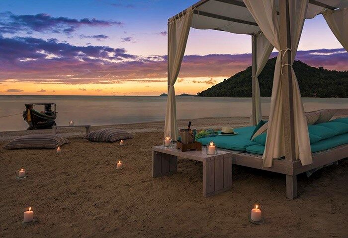 Beachfront canopy bed at sunset, Avani+ Samui Resort, a gay-friendly hotel. Candles illuminate the sand near a boat. Romantic getaway.
