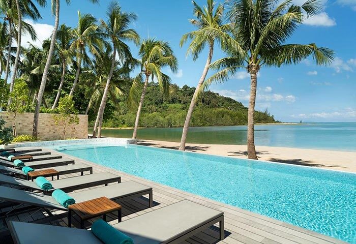 Avani+ Samui Resort's pool, a gay-friendly hotel. Palm trees line the beach. Lounge chairs await guests at this tropical paradise.