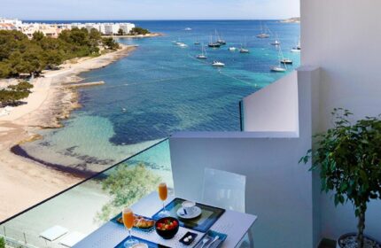 Balcony view at AxelBeach Ibiza Suites, a gay-friendly, adults-only hotel. Breakfast table overlooks the beach and boats. Spa and beach club visible.