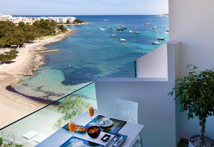 Balcony view at AxelBeach Ibiza Suites, a gay-friendly, adults-only hotel. Breakfast table overlooks the beach and boats. Spa and beach club visible.