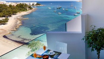 Balcony view at AxelBeach Ibiza Suites, a gay-friendly, adults-only hotel. Breakfast table overlooks the beach and boats. Spa and beach club visible.