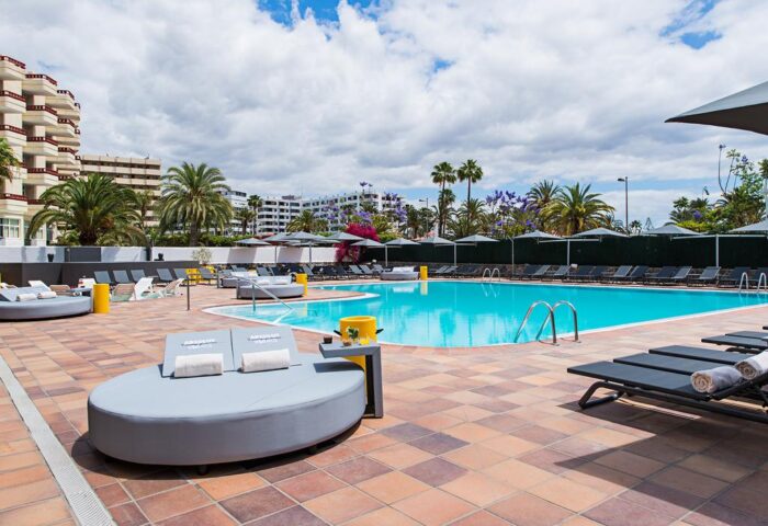 Pool area at AxelBeach Maspalomas, a gay-friendly, adults-only apart-lounge club in Gran Canaria. Sun loungers, palm trees, blue sky.