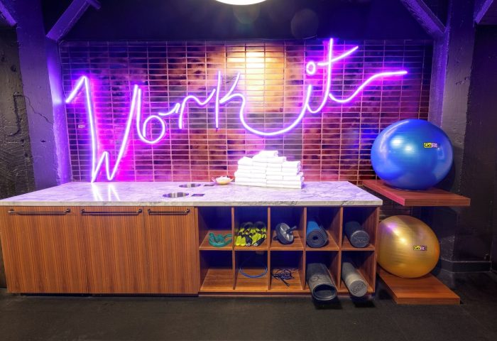 Axiom Hotel's gym. Work it neon sign above towels, yoga mats, and exercise balls. Gay-friendly hotel fitness center.