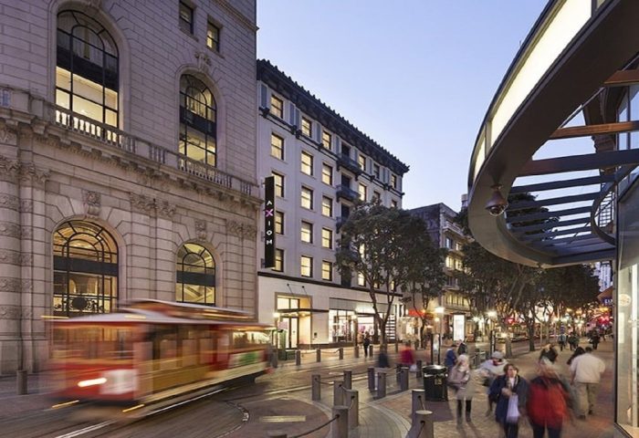 Exterior of the Gay-Friendly Axiom Hotel. A streetcar passes by on the street. People stroll along the sidewalk in front of the hotel.