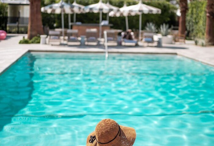 Woman in hat at Azure Sky Hotel adults-only pool. Palm Springs gay-friendly hotel with mountain views, striped umbrellas, and turquoise water.