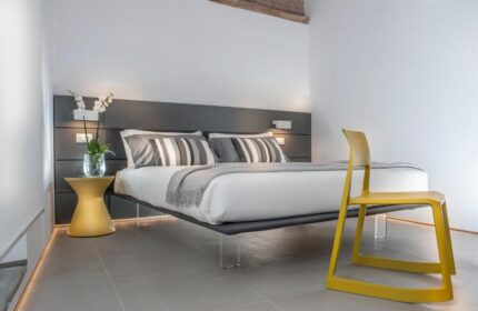 Gay-friendly hotel B&B second floor room. Bed with striped pillows, yellow chair, bedside table with flowers. Modern design, clean lines.