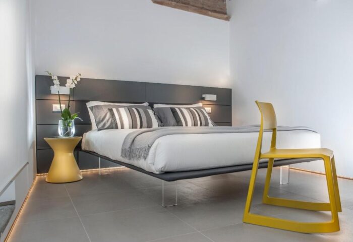 Gay-friendly hotel B&B second floor room. Bed with striped pillows, yellow chair, bedside table with flowers. Modern design, clean lines.