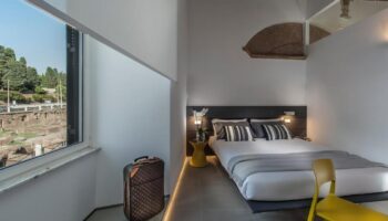 B&B First Floor room: Gay-friendly hotel bedroom with double bed, yellow chair, suitcase, and view of Roman ruins. Modern, stylish design.
