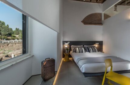B&B First Floor room: Gay-friendly hotel bedroom with double bed, yellow chair, suitcase, and view of Roman ruins. Modern, stylish design.