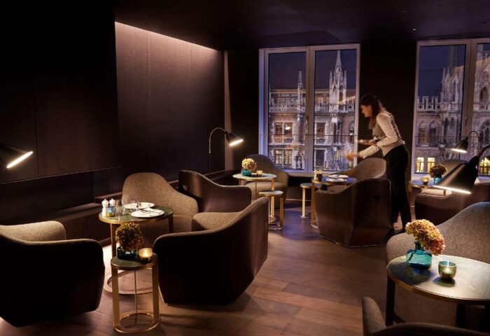 BEYOND by Geisel gay-friendly hotel lounge. Stylish seating, dark walls, and a server. Munich city view from the window. Upscale design.