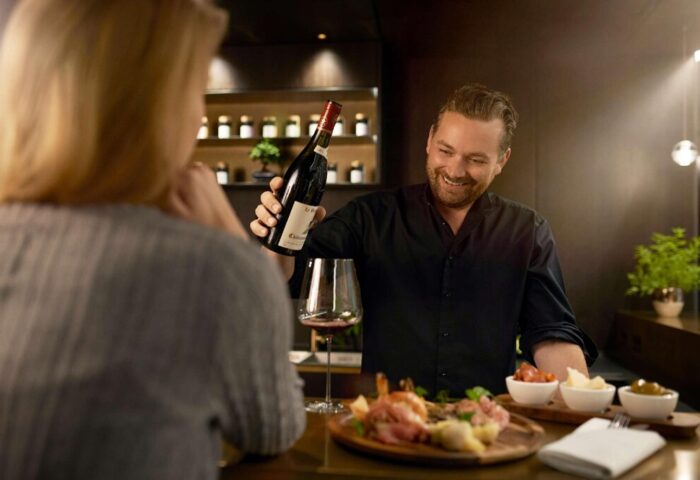 BEYOND by Geisel, a gay-friendly hotel. Waiter pours wine for a guest. Charcuterie board with cheese, olives, and tomatoes on the bar.