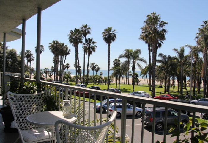 Balcony view at Bayside Hotel, a gay-friendly hotel. Palm trees lead to the beach. A table and chairs sit on the balcony. Cars parked below.