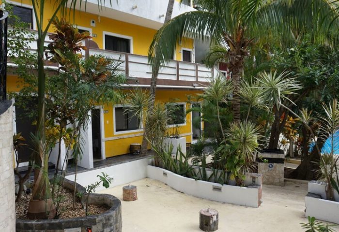 Beach Energy Hotel By BFH, a gay-friendly hotel. Exterior view of the yellow building surrounded by tropical plants and a glimpse of the pool.