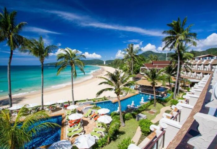 Gay-friendly Beyond Resort Karon, Thailand. Beachfront view with palm trees, pools, and clear blue sky. Ideal gay travel destination.