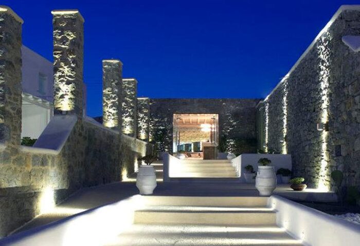 Bill & Coo Suites and Lounge entrance at night. Luxury gay-friendly hotel in Mykonos, Greece. Stone walls, illuminated steps, and elegant design.