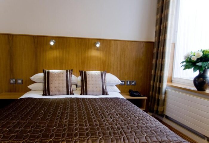 Blooms Hotel bedroom. Gay-friendly hotel room with a double bed, brown patterned quilt, pillows, bedside tables, and a vase of flowers by the window.