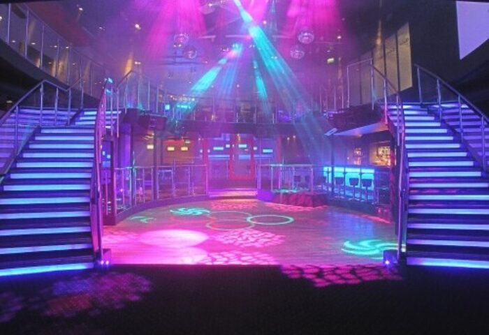 Blooms Hotel gay-friendly bar/club interior. Stairs lead to a dance floor with colorful lights. A bar area is visible in the background.