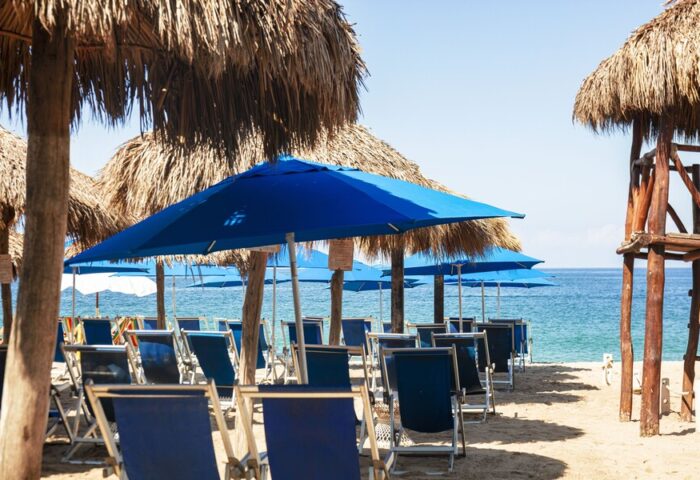Beach at Blue Chairs Resort by the Sea, an adults-only, gay-friendly hotel. Blue umbrellas and chairs line the sand, facing the ocean.