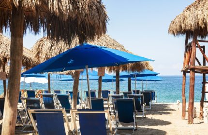 Beach at Blue Chairs Resort by the Sea, an adults-only, gay-friendly hotel. Blue umbrellas and chairs line the sand, facing the ocean.