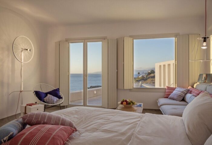 Boheme Mykonos Hotel adults-only room. Gay-friendly hotel with sea view. Stylish interior with bed, chair, and windows overlooking the Aegean Sea.