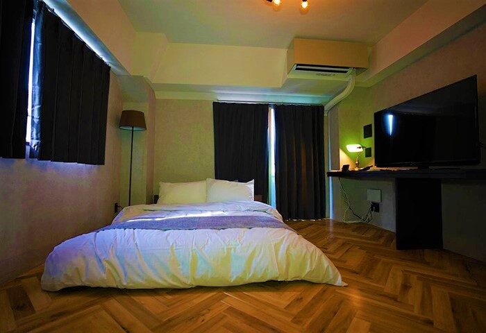 Bedroom at Book Tea Bed SHINJUKU-GYOEN, a gay-friendly hotel. Bed with white linens, dark curtains, and a TV. Wooden floor. Comfy stay.