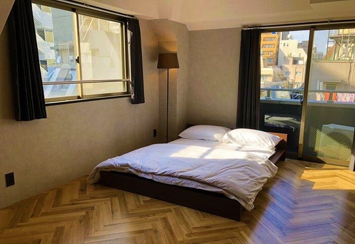 Book Tea Bed SHINJUKU-GYOEN gay-friendly hotel room. Bed with white linens, herringbone floor, windows with curtains, floor lamp, balcony view.