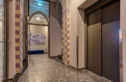 Boutique Hotel 026 Köln St. Michael interior. Hallway view with elevator and arched doorway. Gay-friendly hotel with unique design elements.