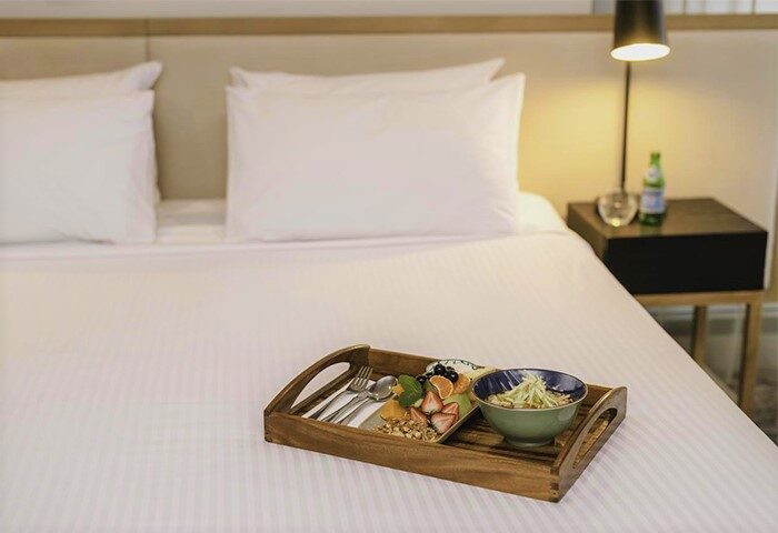 Brady Hotels Central Melbourne gay-friendly hotel room service. Tray with fruit, nuts, and noodles on a bed. Lamp and nightstand visible.