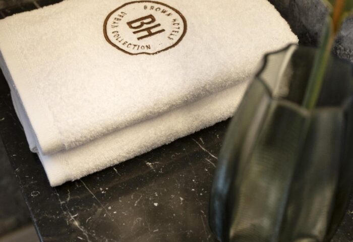 Brown Acropol Hotel detail: Stack of white towels with the Brown Hotels logo, vase, and glasses on a marble counter. Gay-friendly hotel.