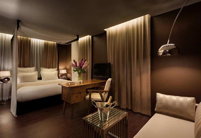 Brown TLV Hotel room. Gay-friendly hotel in Tel Aviv features a bed with curtains, a desk with flowers, a TV, and a seating area with champagne.