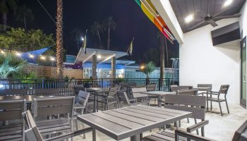 Outdoor dining area at CCBC Resort Hotel, a gay men's resort. Tables and chairs on a patio, palm trees, and a rainbow awning. Gay-friendly hotel.