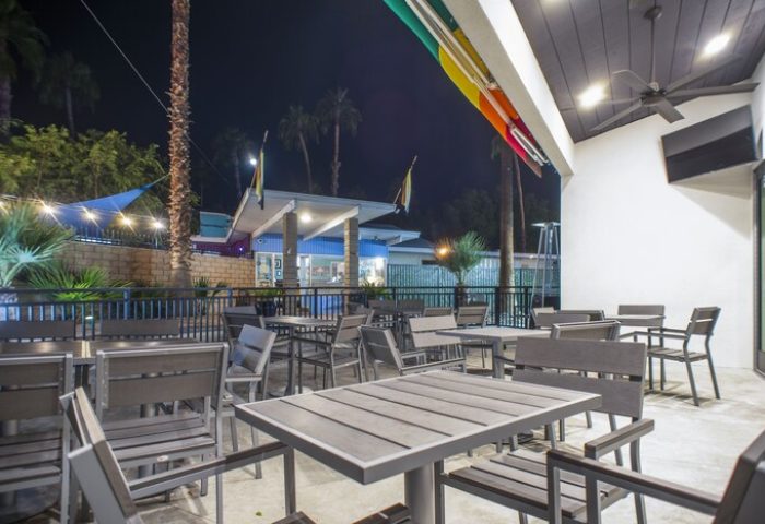Outdoor dining area at CCBC Resort Hotel, a gay men's resort. Tables and chairs on a patio, palm trees, and a rainbow awning. Gay-friendly hotel.