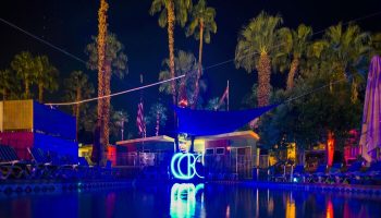 CCBC Resort Hotel pool at night. Gay men's resort with palm trees, lounge chairs, and neon CCBC logo reflected in the water. Gay-friendly hotel.