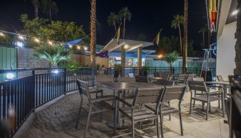 Outdoor dining area at CCBC Resort Hotel, a gay men's resort. Tables and chairs are set on a patio under palm trees at night. Gay-friendly hotel.