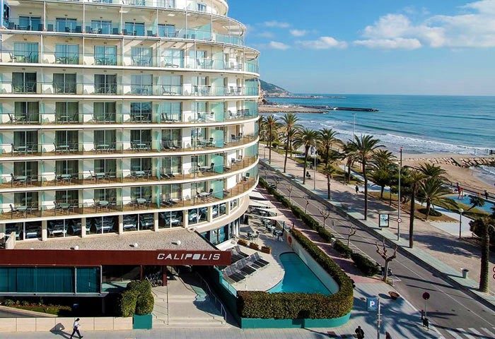 Gay-Friendly Hotel Calipolis view. Beachfront hotel with balconies overlooking the Mediterranean Sea. Palm trees line the coast. Gay travel.