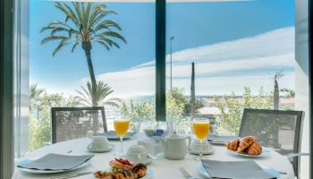Breakfast at Gay-Friendly Hotel Calipolis. Table set with pastries, juice, & coffee. Ocean view with palm trees. Sitges, Spain.