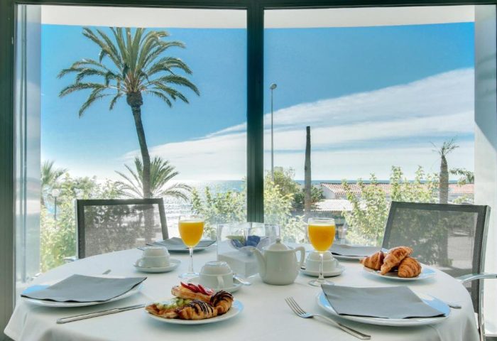 Breakfast at Gay-Friendly Hotel Calipolis. Table set with pastries, juice, & coffee. Ocean view with palm trees. Sitges, Spain.