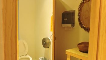 Bathroom interior at a gay-friendly capsule hostel. Toilet, sink, mirror, and trash can visible. Budget travel lodging option.