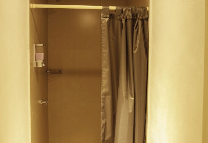 Shower stall in a capsule hostel. Gray curtain, dispenser, & shower head. Gay-friendly hotel bathroom. Budget travel for gay men.
