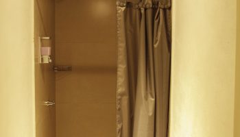 Shower stall in a capsule hostel. Gray curtain, dispenser, & shower head. Gay-friendly hotel bathroom. Budget travel for gay men.