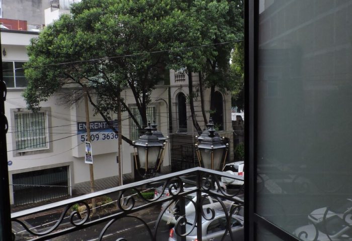 View from a gay-friendly capsule hostel balcony. Street scene with cars, trees, and buildings. Travel lodging for gay men.