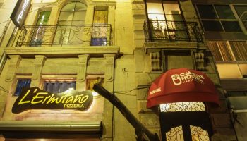 Capsule Hostel, a gay-friendly hotel. Exterior view at night with a red awning. Pizzeria L'Ermitaño next door. Balconies with iron railings.