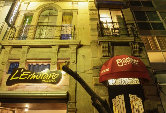 Capsule Hostel, a gay-friendly hotel. Exterior view at night with a red awning. Pizzeria L'Ermitaño next door. Balconies with iron railings.